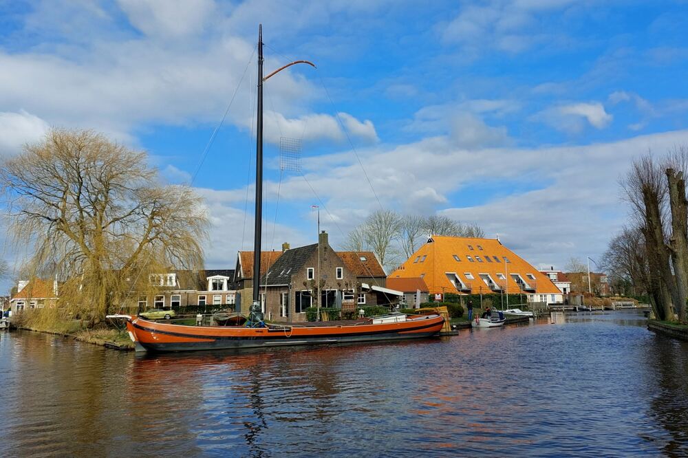 Historisch zeilschip afgemeerd aan het water in Heeg, met dorpshuizen en een groot oranje pannendak op de achtergrond